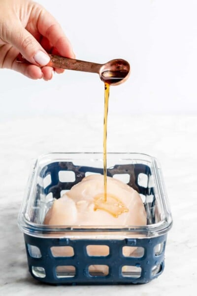 Hand holding a measuring spoon and drizzling sesame oil over a chicken breast in a glass container with a blue netted silicone liner.