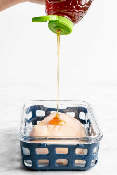 Hand squeezing honey over top a chicken breast in a glass container with a blue netted silicone liner.
