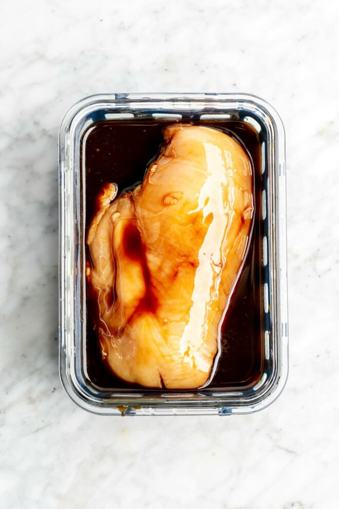 Chicken breast marinating in a dark liquid in a glass container.