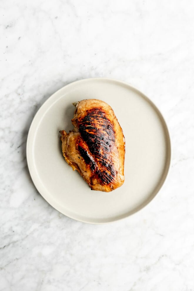 Pan seared teriyaki chicken breast on a round plate on a grey and white marble surface. 