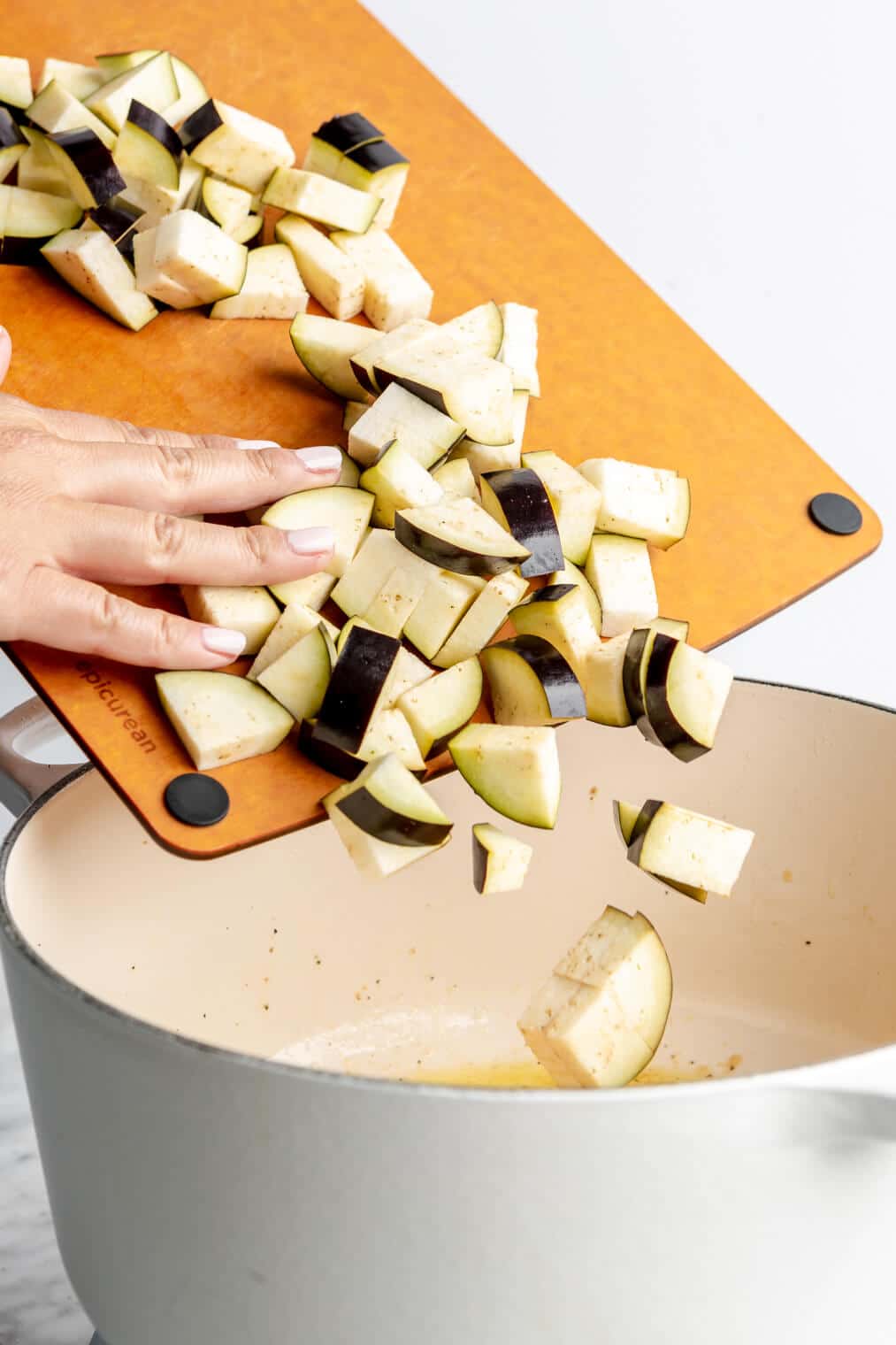 Hand pushing cubed eggplant into a white enameled dutch oven.