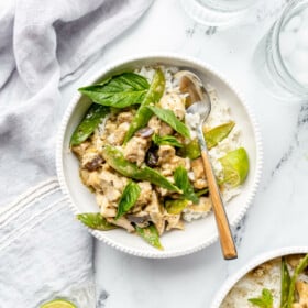 Two bowls of green curry with chicken, eggplant, and snow peas in white bowls with rice. They are sitting on a white and black marble surface with two glasses of ice water and a couple of limes.