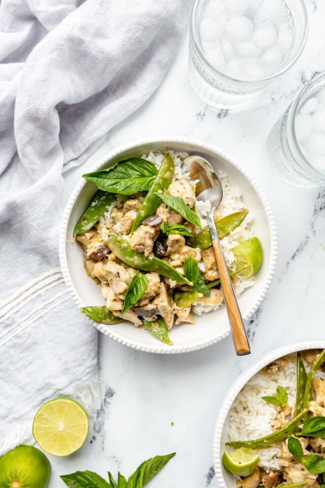 Two bowls of green curry with chicken, eggplant, and snow peas in white bowls with rice. They are sitting on a white and black marble surface with two glasses of ice water and a couple of limes. 
