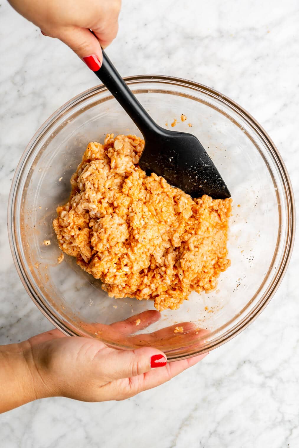 Hand holding a spatula stirring together a ground chicken mixture in a glass bowl.