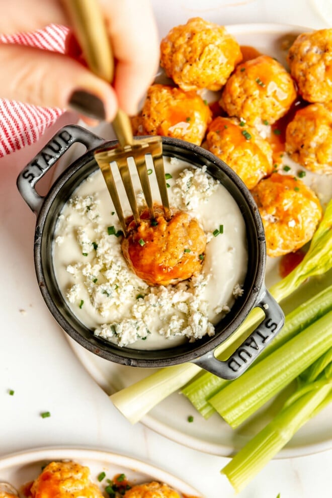 Hand holding a gold fork with a buffalo meatball on the end. The meatball is being dipped into a small dutch oven with a blue cheese dipping sauce. 