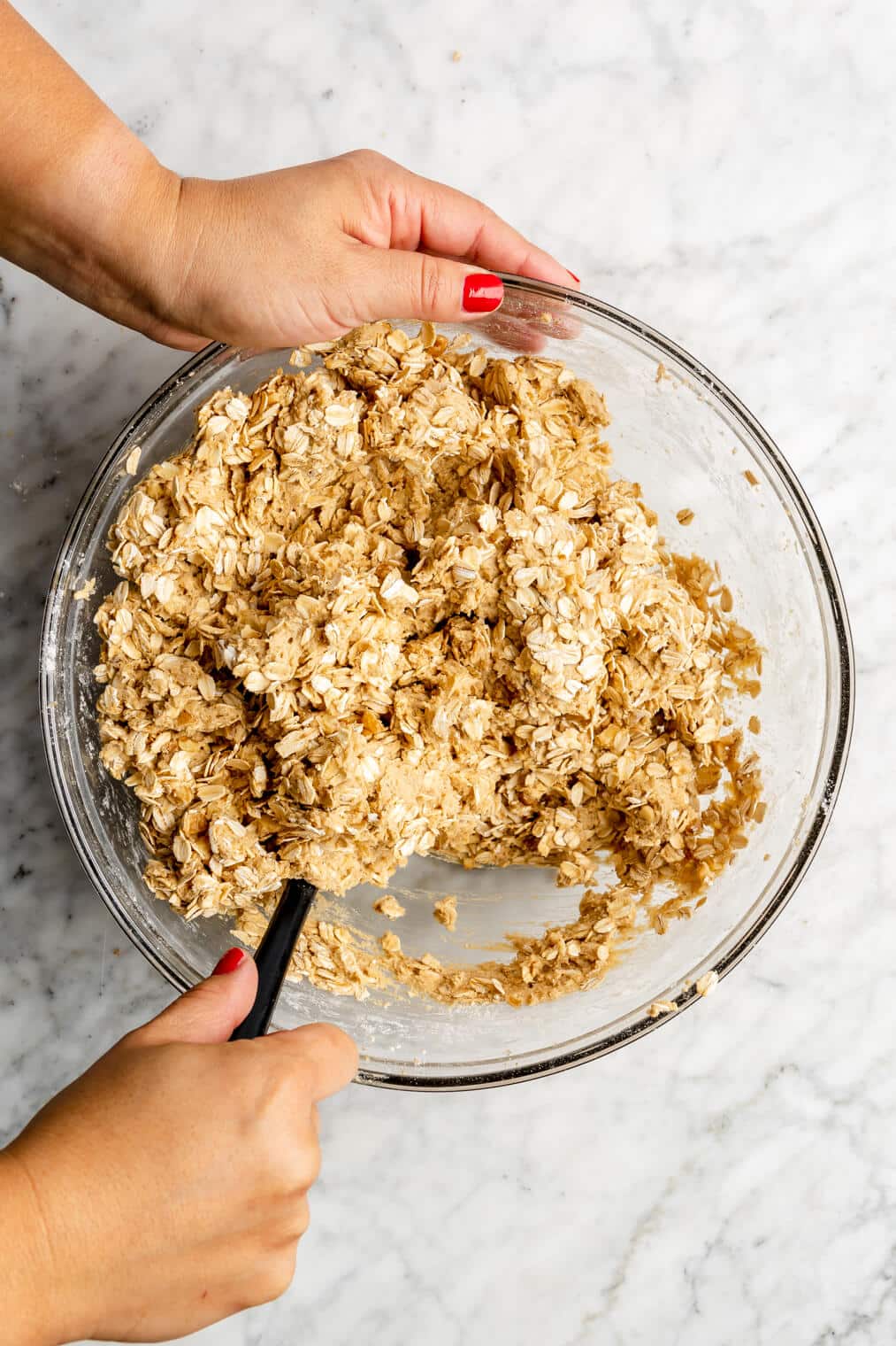 Hand holding spatula stirring oats into cookie dough mixture.