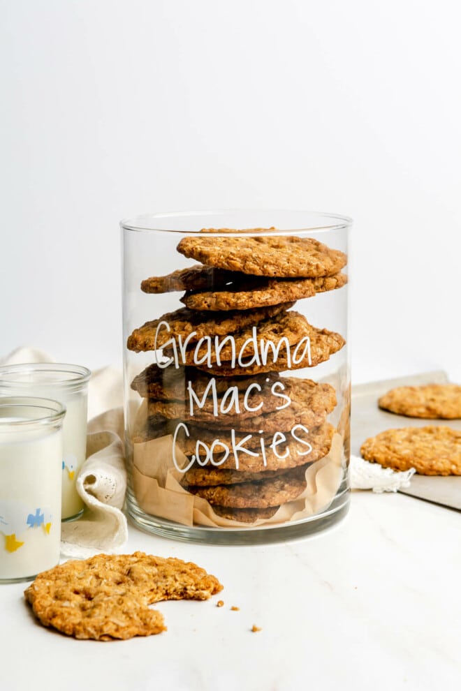 A clear, glass cookie jar with the words "Grandma Mac's Cookies" written on the front is filled with oatmeal cookies. There is also a bit taken out of a cookie on the table in front of the cookie jar.