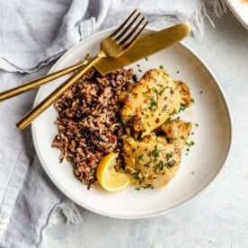 A white plate of wild rice and lemon garlic chicken thighs. Also on the plate: a squeezed lemon wedge and a gold fork and knife