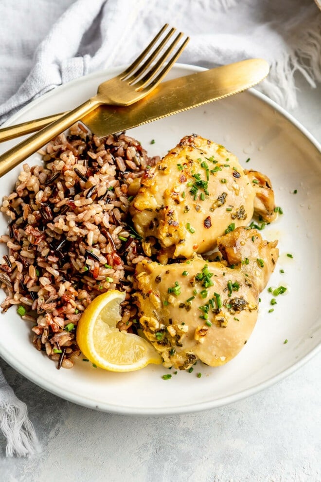 A white plate of wild rice and lemon garlic chicken thighs. Also on the plate: a squeezed lemon wedge and a gold fork and knife