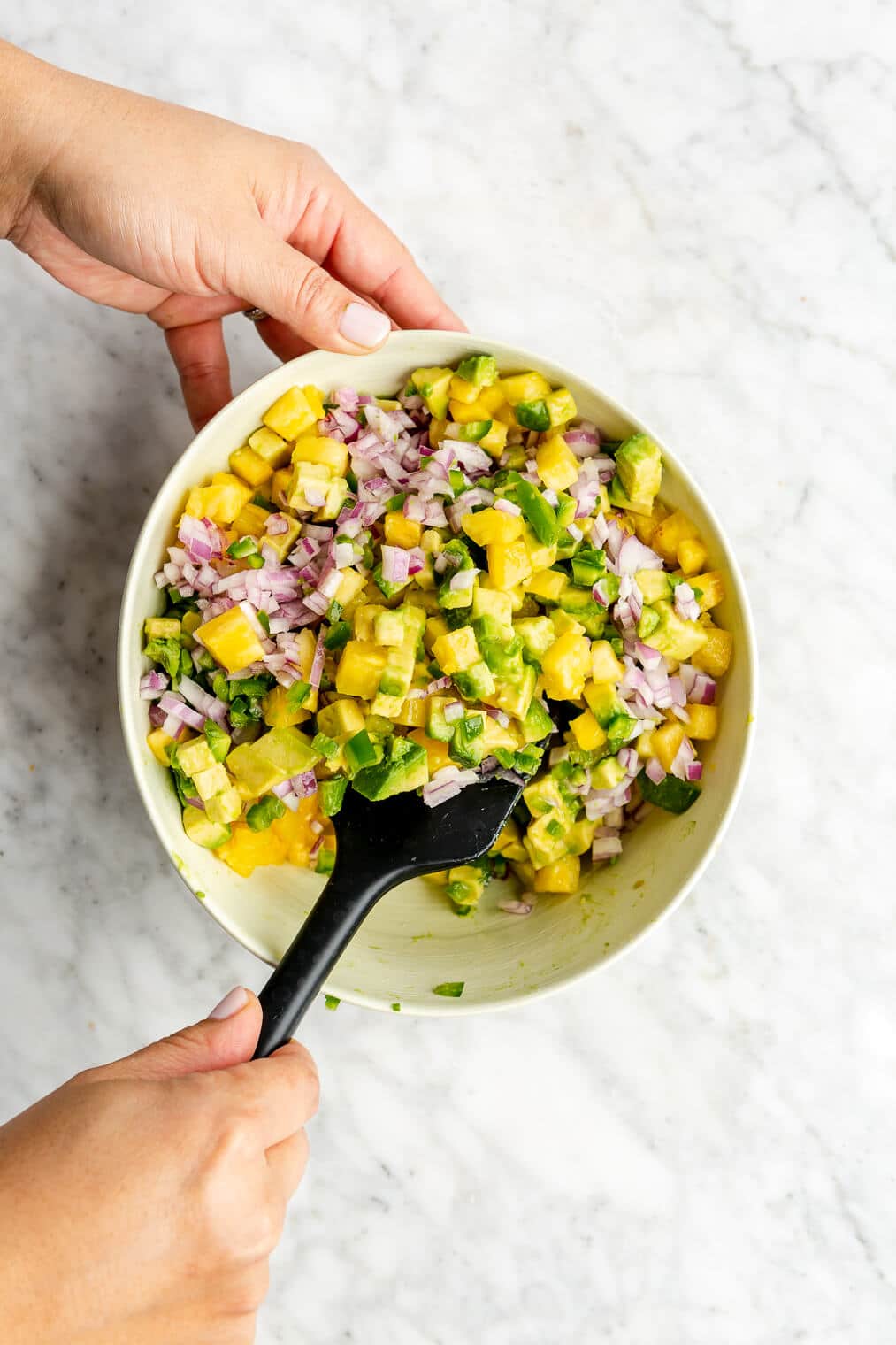 Hand tossing pineapple salsa ingredients in a bowl with a black spatula.