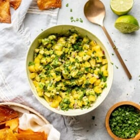 A white bowl of pineapple salsa on a grey and white marble surface. There are fresh tortilla chips to the bottom left and sprinkled near the top. There is also a copper spoon to the top right and a wooden bowl of cilantro to the bottom right.