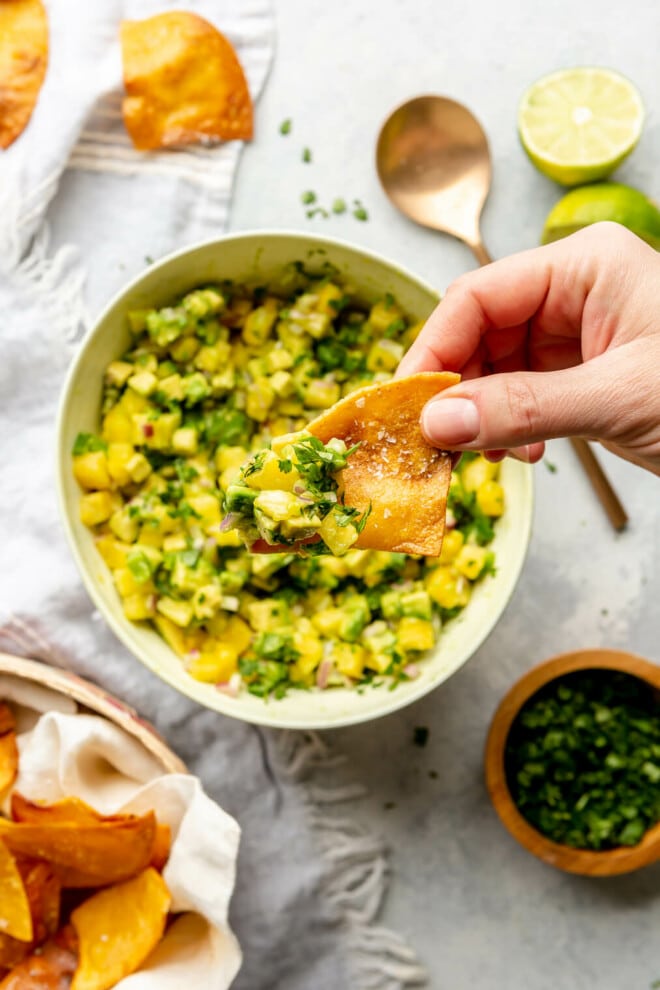 Hand holding a fresh tortilla chip dipped in a bowl of pineapple salsa. There is a basket of fresh chips in the bottom left corner and a small wooden bowl of chopped cilantro to the bottom right. 