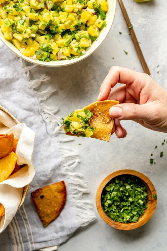 Hand holding a fresh tortilla chip over a grey surface. There is a bowl of pineapple salsa to the top and a small wooden bowl of chopped cilantro to the bottom right. 