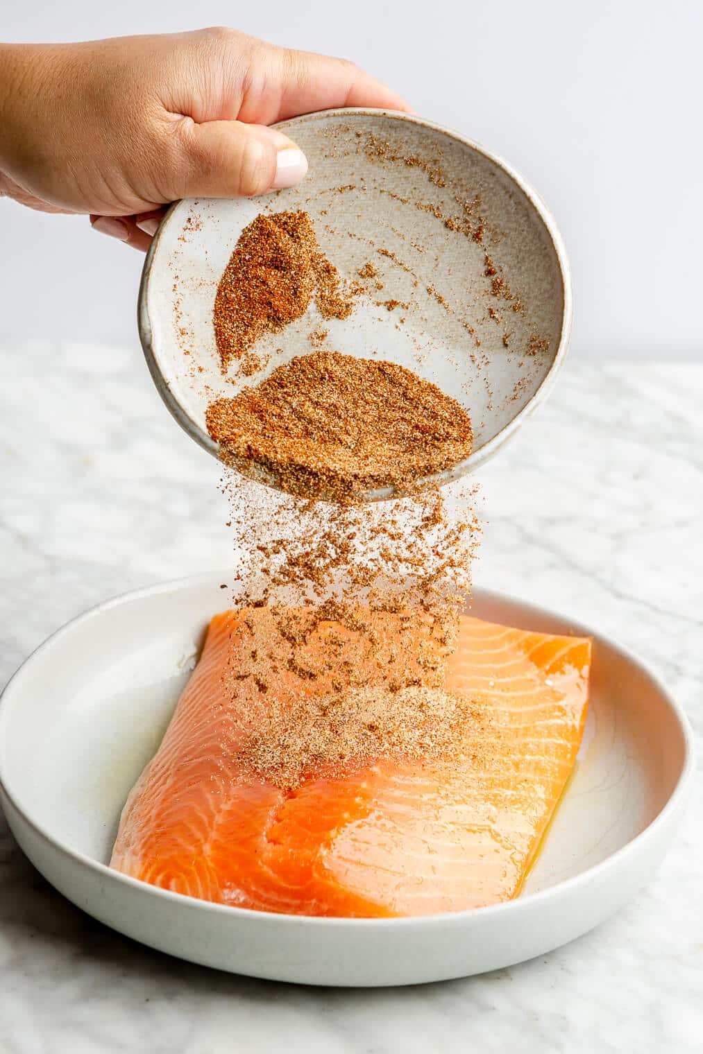 Hand pouring a mixture of spices over top an oil coated salmon filet.