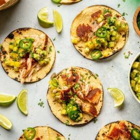 Grey tabletop with salmon tacos on charred corn tortillas garnished with pineapple salsa, jalapeno, and cilantro.