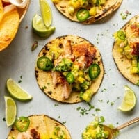 Grey tabletop with three salmon tacos on charred corn tortillas garnished with pineapple salsa, jalapeno, and cilantro.