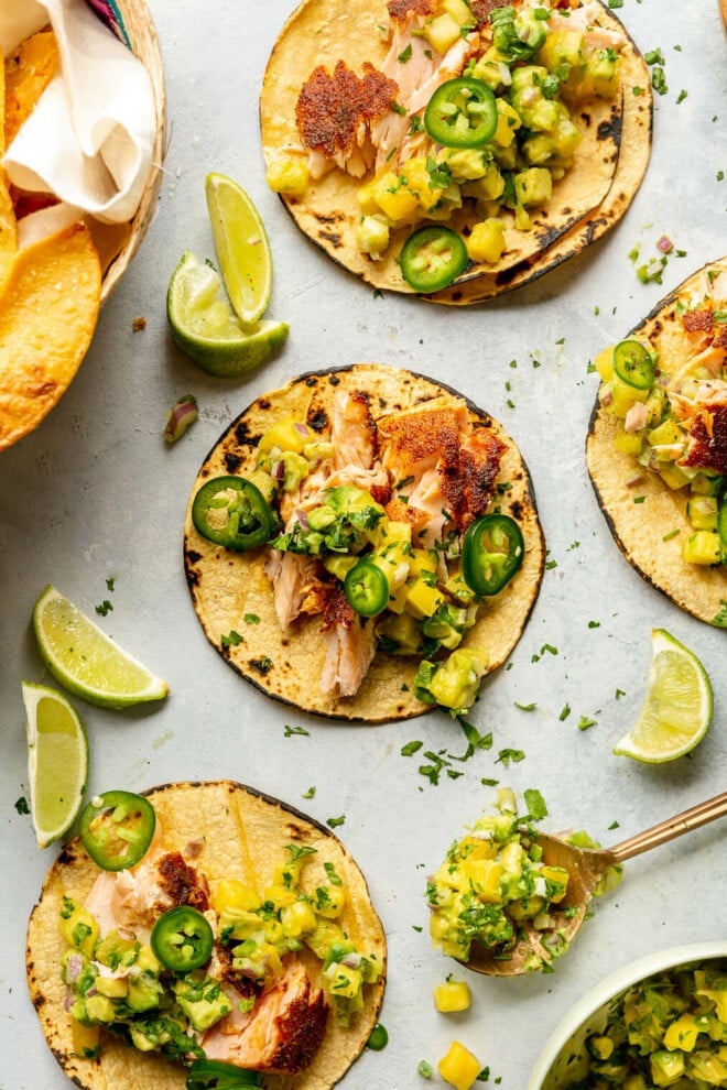 Grey tabletop with three salmon tacos on charred corn tortillas garnished with pineapple salsa, jalapeno, and cilantro.