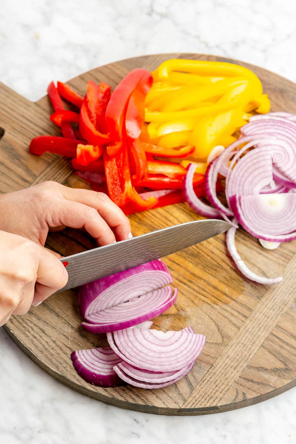 A person using a large knife to slice a red onion on a wood cutting board. Also on the cutting board: sliced yellow and red bell peppers.