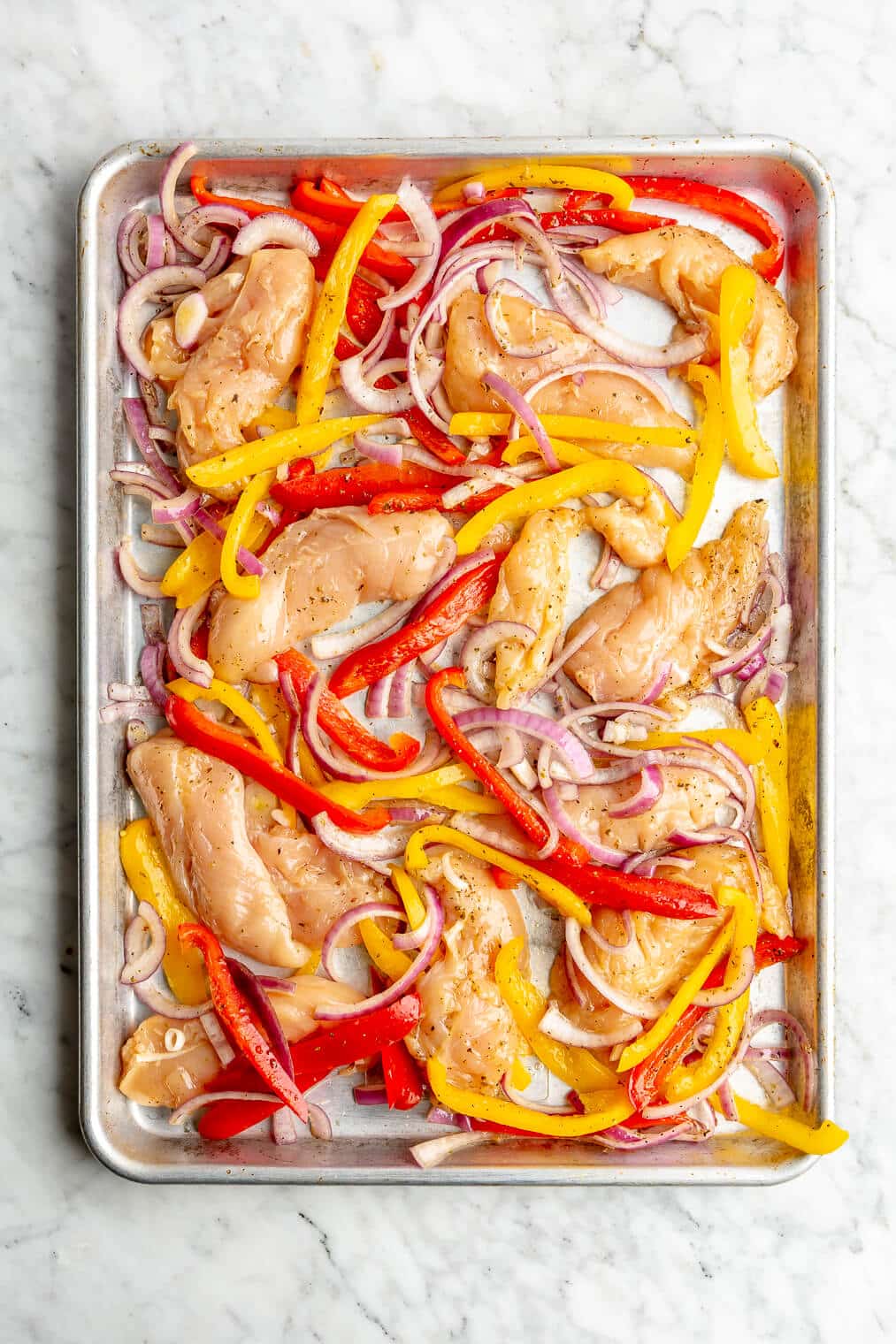 A sheet pan of marinated raw chicken tenders, bell peppers, and onion.