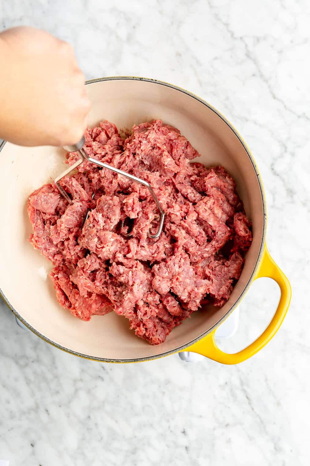 Ground beef being cooked and mashed with a metal potato masher in a yellow enameled cast iron pot.