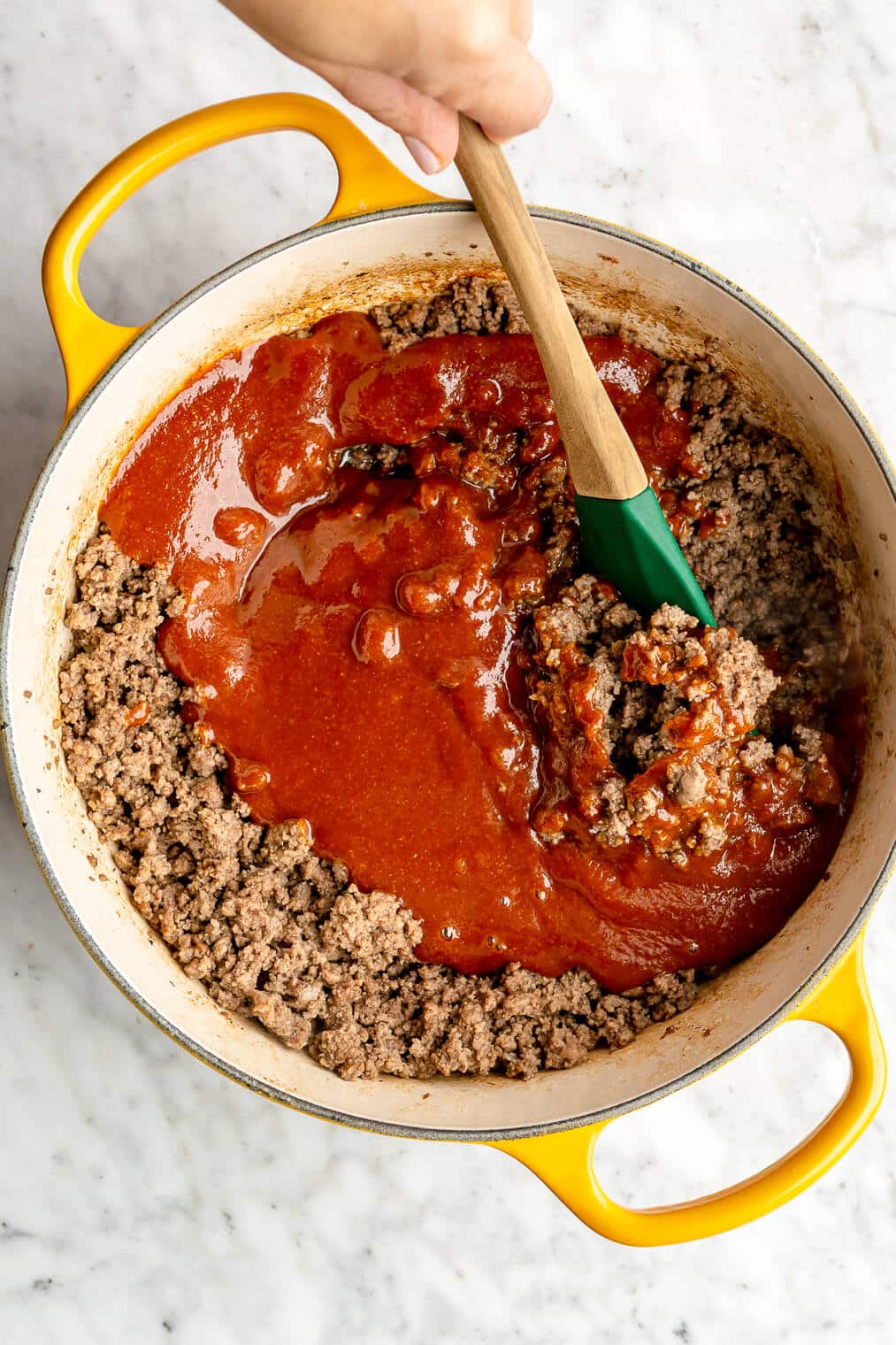 Sloppy joe sauce being mixed into cooked ground beef in a yellow enameled cast iron pot.