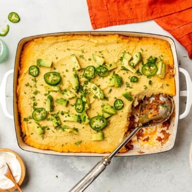Top down view of tamale pie in a casserole dish on a grey surface. There is a scoop out of the casserole. It's topped with slices of avocado and jalapeno.