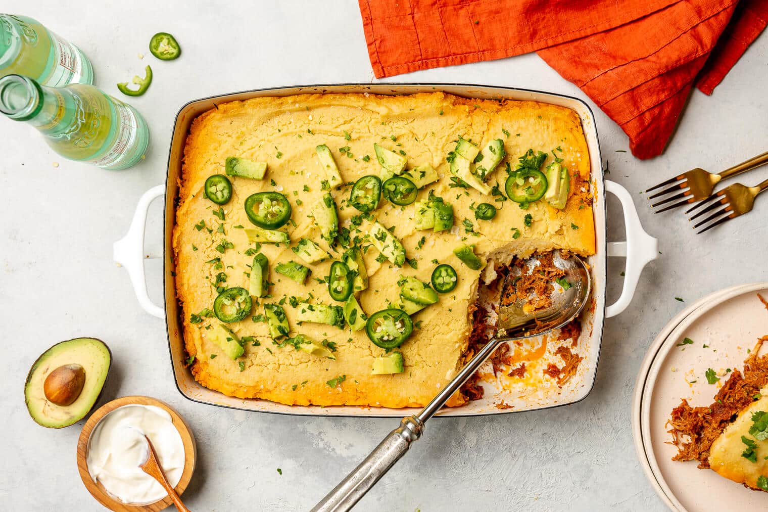 Top down view of tamale pie in a casserole dish on a grey surface. There is a scoop out of the casserole. It's topped with slices of avocado and jalapeno.