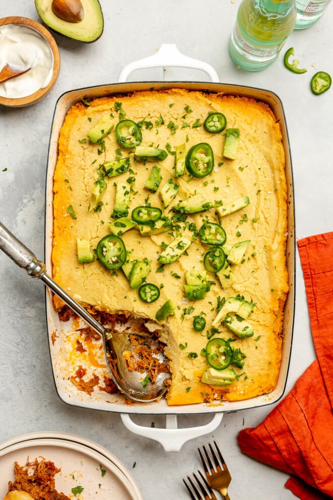 Top down view of tamale pie in a casserole dish on a grey surface. There is a scoop out of the casserole. It's topped with slices of avocado and jalapeno. 