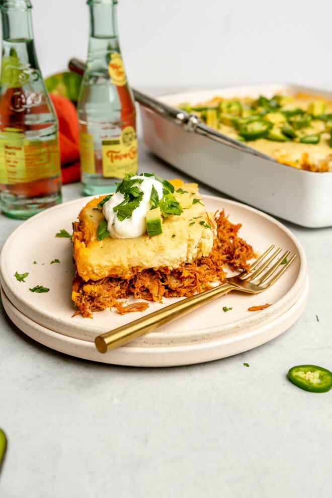 Slice of tamale pie on a stack of two light pink plates. There are a couple bottles of Topo Chico sparkling water in the background and a casserole dish. The slice of pie is topped with a dollop of sour cream and a sprinkle of cilantro.