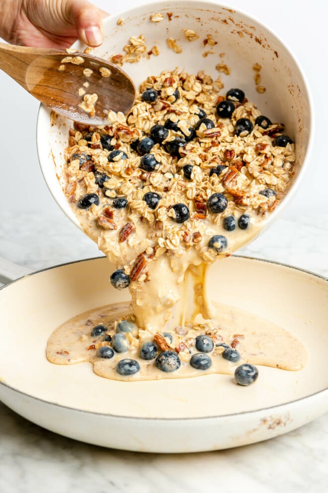 Hand holding a white bowl and pouring blueberry oatmeal bake ingredients into a white skillet using a wooden spoon.
