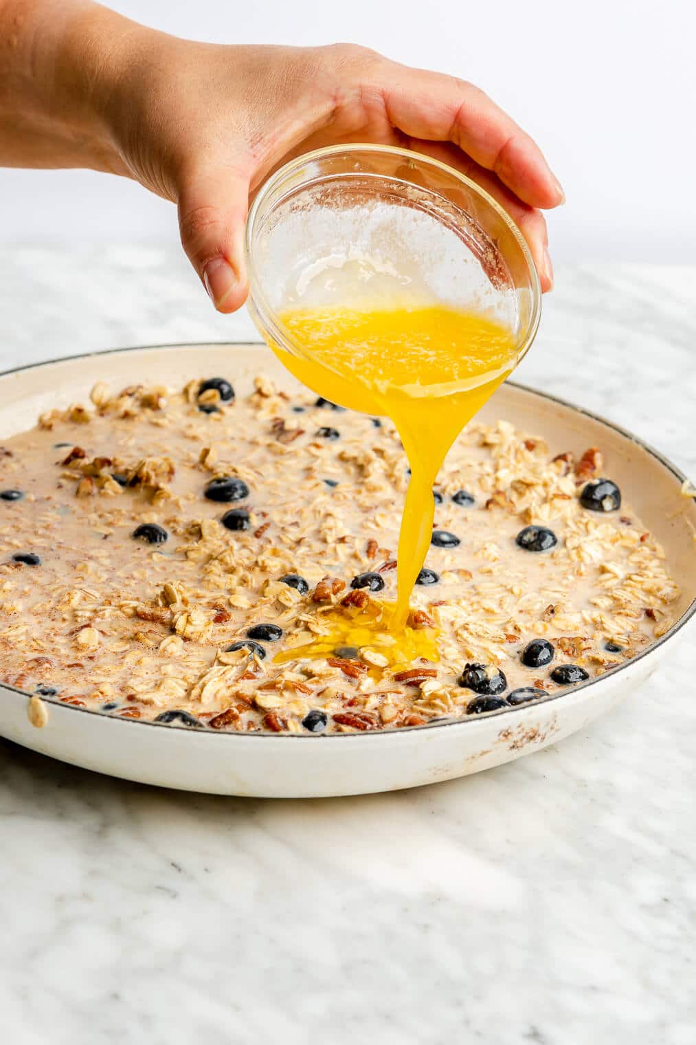 Hand drizzling melted butter on top of blueberry oatmeal ingredients in a white skillet.