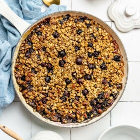 Top down view of baked blueberry oatmeal in a round skillet. The skillet is sitting on a white tile surface. There is a blue linen draped to the bottom, a couple of gold spoons, a bowl of blueberries, and a white pot of honey with a honeycomb serving utensil resting on top.