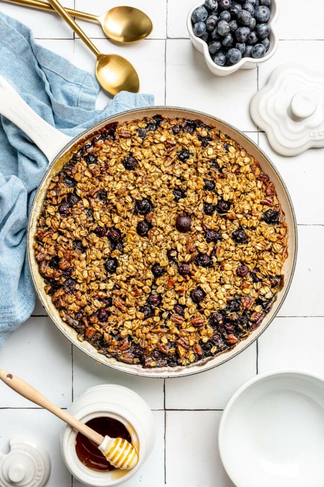 Top down view of baked blueberry oatmeal in a round skillet. The skillet is sitting on a white tile surface. There is a blue linen draped to the bottom, a couple of gold spoons, a bowl of blueberries, and a white pot of honey with a honeycomb serving utensil resting on top.