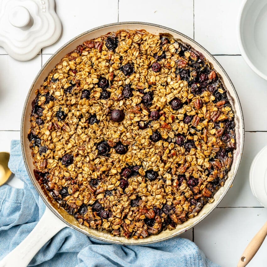 Top down view of baked blueberry oatmeal in a round skillet. The skillet is sitting on a white tile surface. There is a blue linen draped to the bottom, a couple of gold spoons, a bowl of blueberries, and a white pot of honey with a honeycomb serving utensil resting on top.