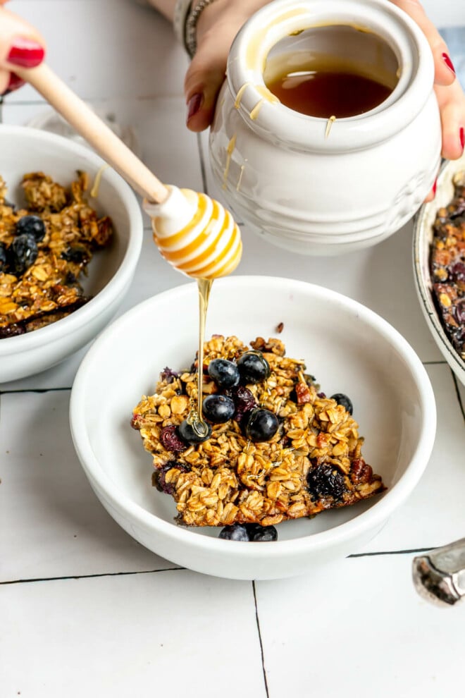 A white bowl with a serving of blueberry baked oats in the bowl. A hand is drizzling honey over the bowl. 