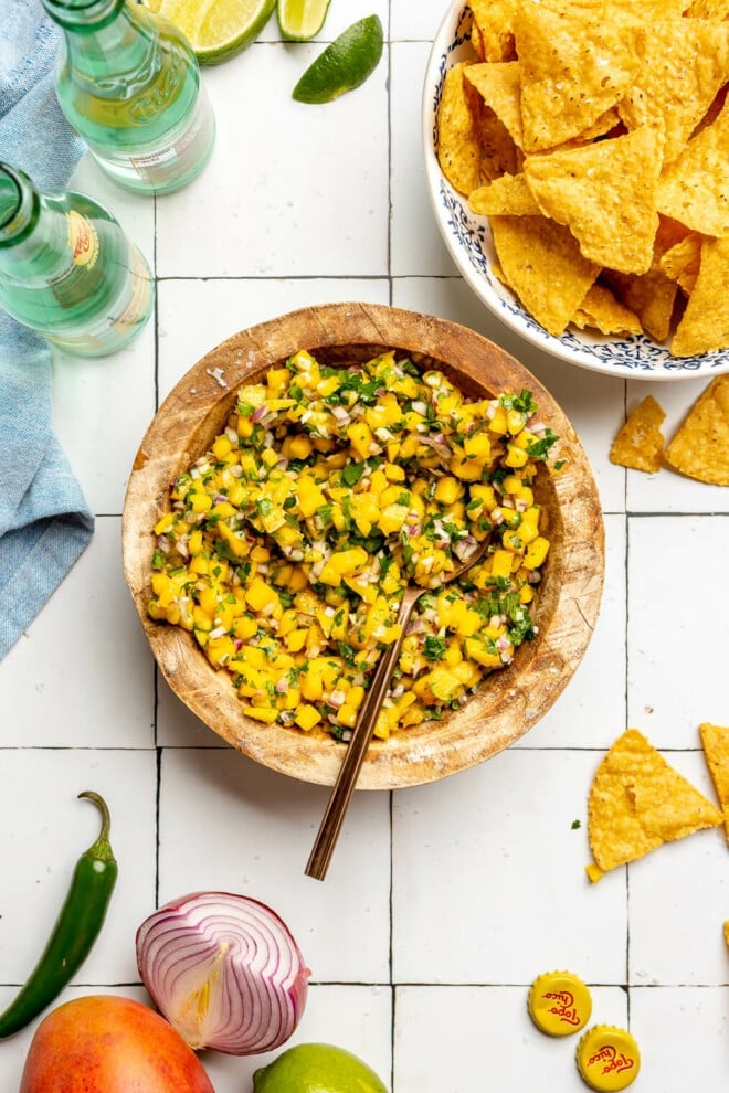 A wooden bowl of finely diced homemade mango salsa sitting on a white tiled table next to a bowl of tortilla chips, a Topo Chico sparkling water, a mango, half of an onion, a lime, and a serrano pepper.