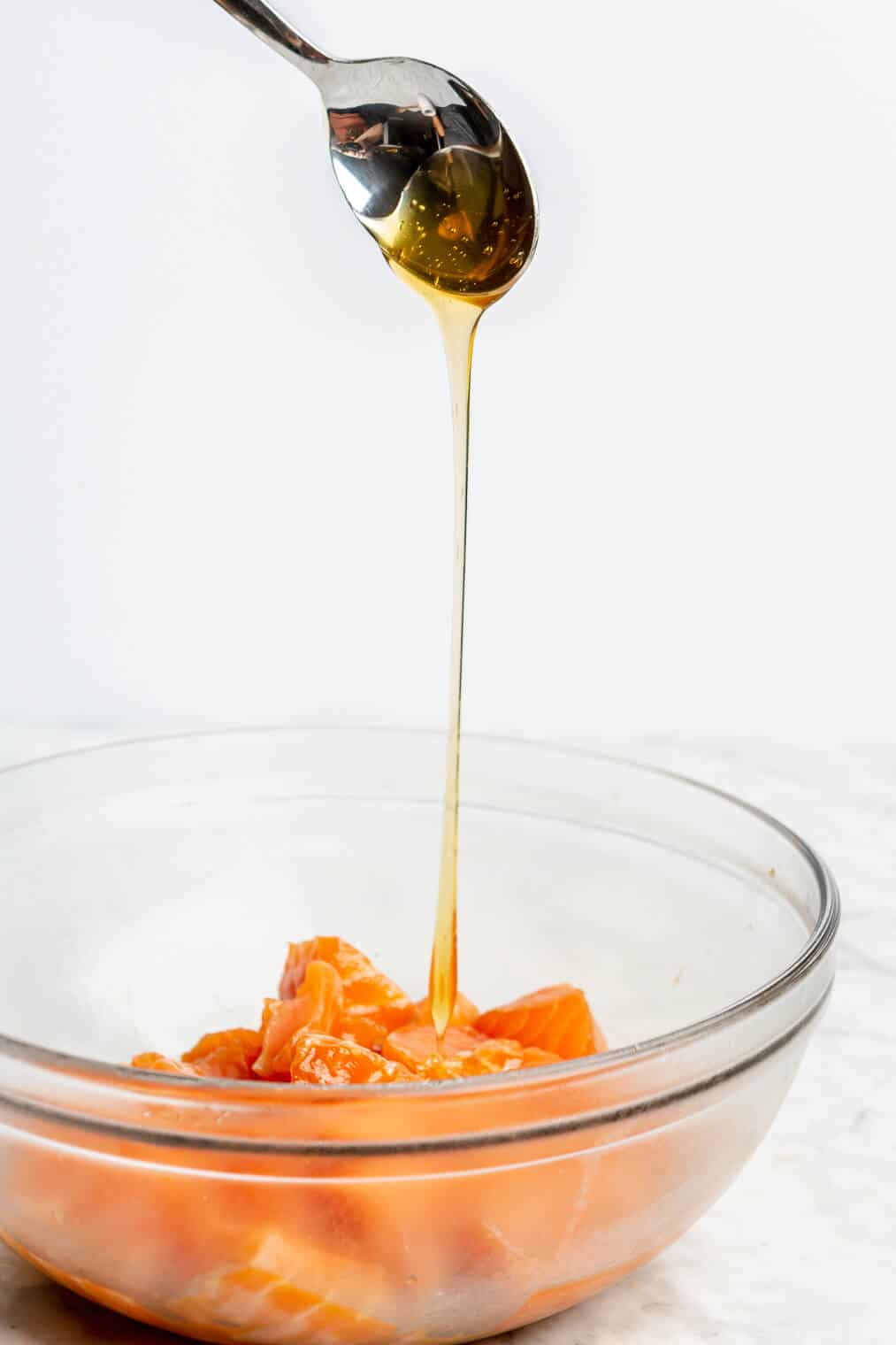 Honey being drizzled over a clear bowl of cubed, raw salmon.