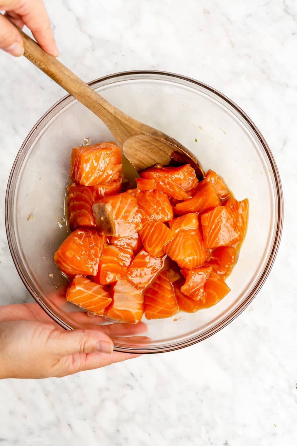A clear bowl of raw, cubed salmon being tossed in honey and teriyaki sauce with a wooden spoon.