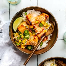 White rice, teriyaki salmon, and mango salsa in a brown bowl sitting on a white tiled table next to a bottle of sparkling water and a white dish towel.