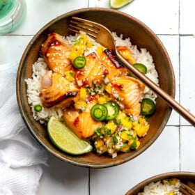 A bowl of white rice topped with teriyaki salmon and mango salsa sitting on a white tiled table next to a white kitchen towel, lime wedges, and bottles of sparkling water.