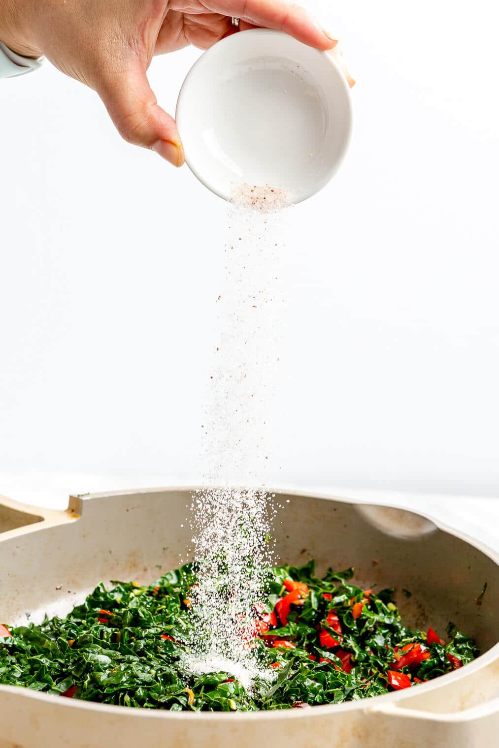 Hand pouring salt from a small white bowl over top kale in a saute pan.