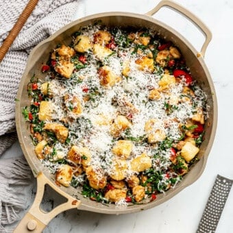 Top down view of a beige pan with gnocchi, sausage, and kale garnished with grated parmesan cheese. There's also a block of parmesan cheese and a microblade to the bottom right and a grey linen with a serving spoon to the top left.