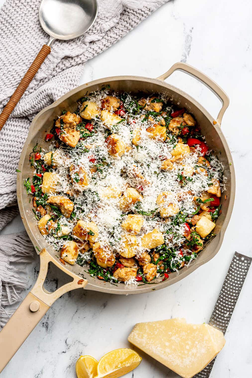 Top down view of a beige pan with gnocchi, sausage, and kale garnished with grated parmesan cheese. There's also a block of parmesan cheese and a microblade to the bottom right and a grey linen with a serving spoon to the top left.