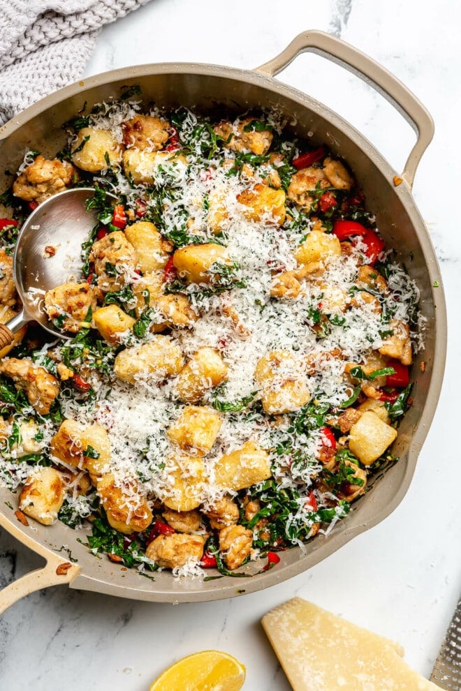 Beige pan with gnocchi, sausage, and kale garnished with parmesan cheese on a white and black marble surface.