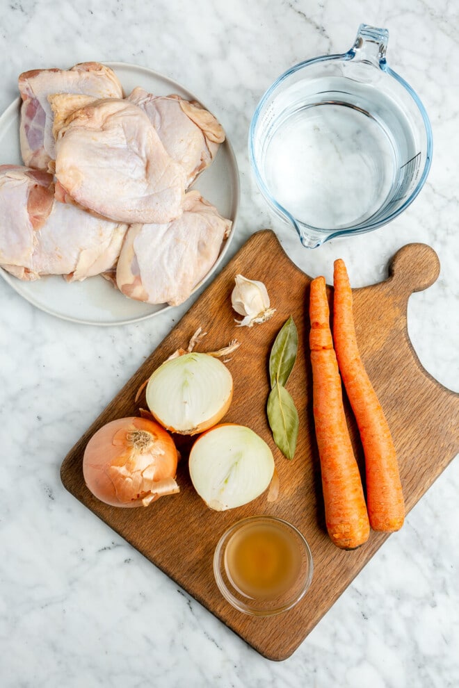 All of the ingredients for homemade chicken broth (bone-in chicken, water, onion, bay leaves, carrots, ACV, and garlic) sitting on marble countertop.