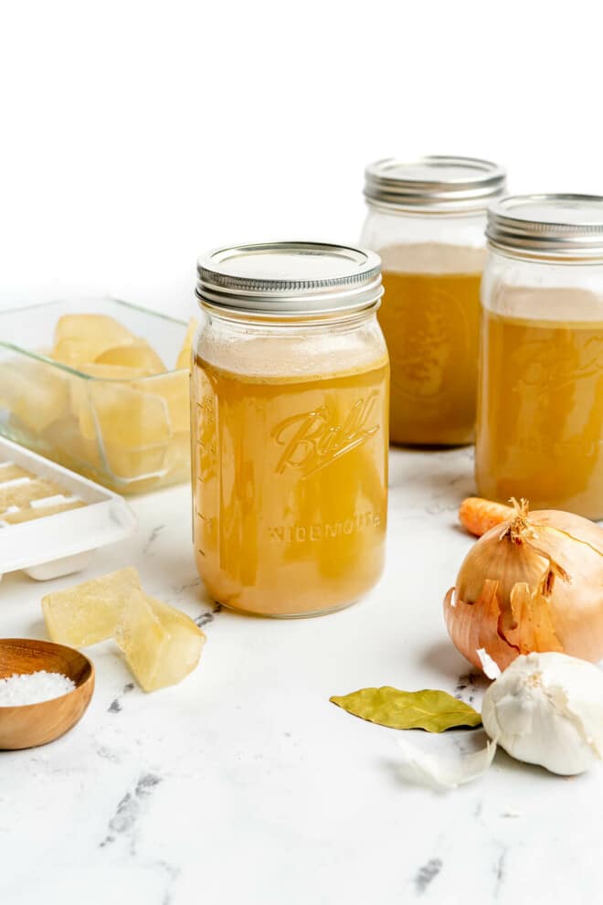 3 large mason jars of homemade chicken broth sitting on a marble countertop next to frozen ice cubes of chicken  broth, an onion, a bulb of garlic, a carrot, and a small bowl of salt.