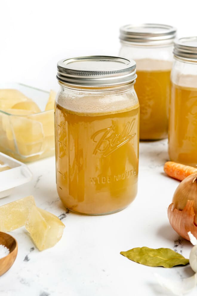 3 large mason jars of homemade chicken broth sitting on a marble countertop next to frozen ice cubes of chicken  broth, an onion, a bulb of garlic, a carrot, and a small bowl of salt.
