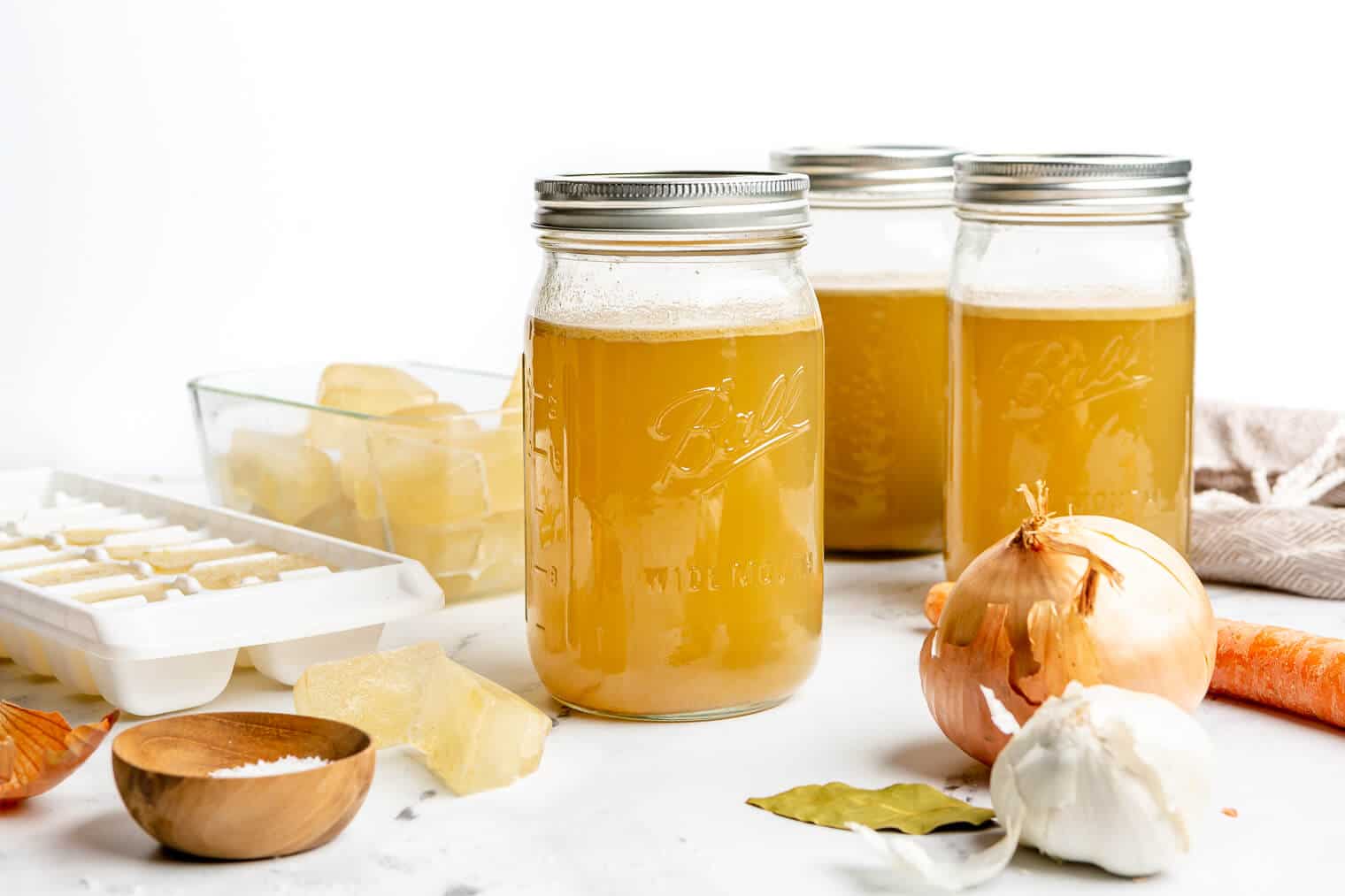 3 large mason jars of homemade chicken broth sitting on a marble countertop next to frozen ice cubes of chicken broth, an onion, a bulb of garlic, a carrot, and a small bowl of salt.