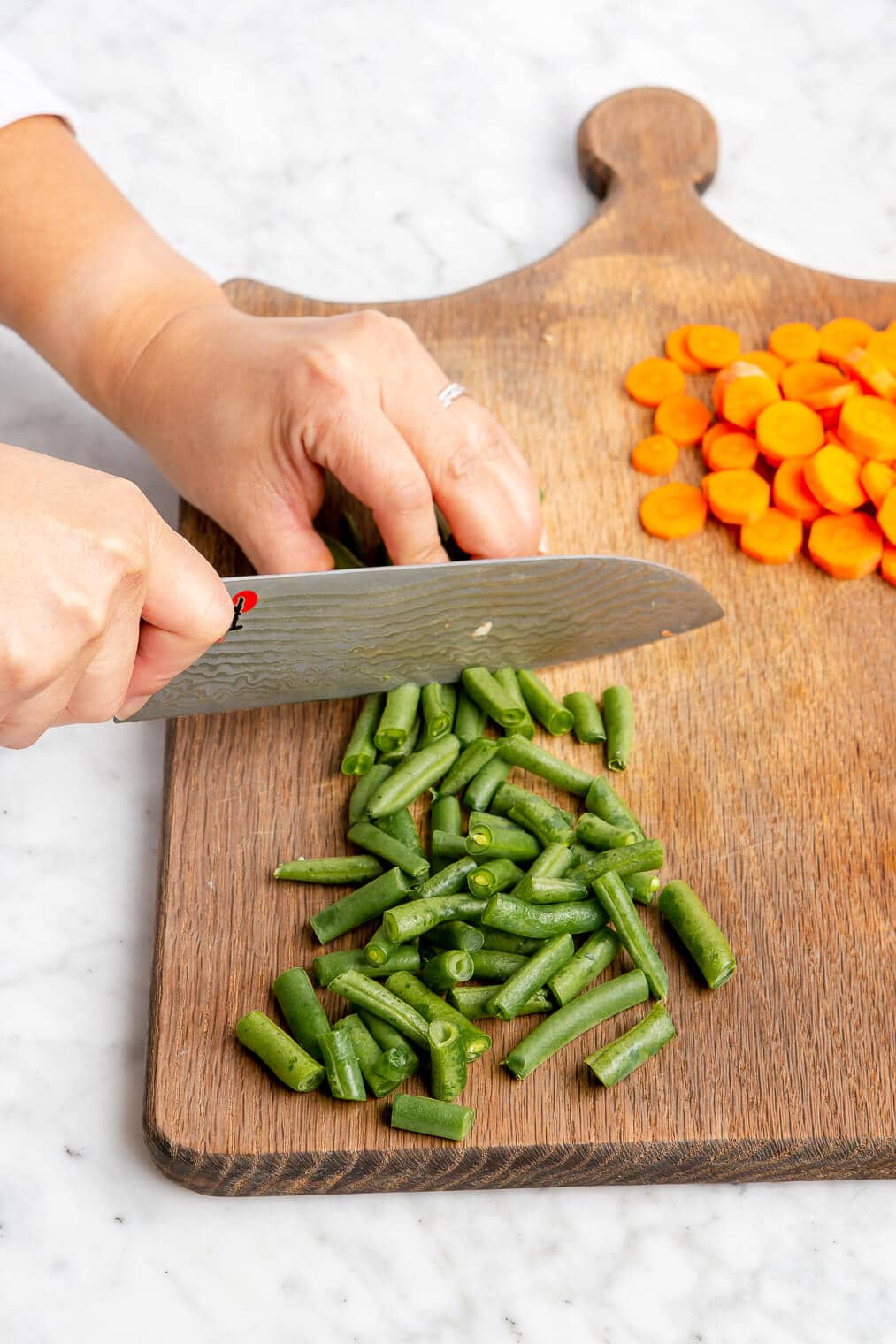 Hand holding a butcher knife to cut green beans. There are sliced carrots in the background on a wooden cutting board.