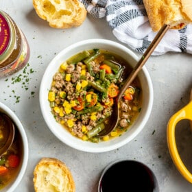 Top down of a white bowl with beef and vegetable soup on a grey surface. There is a jar of bone broth and a yellow dutch oven with soup also on the surface. There are slices of French bread with butter.